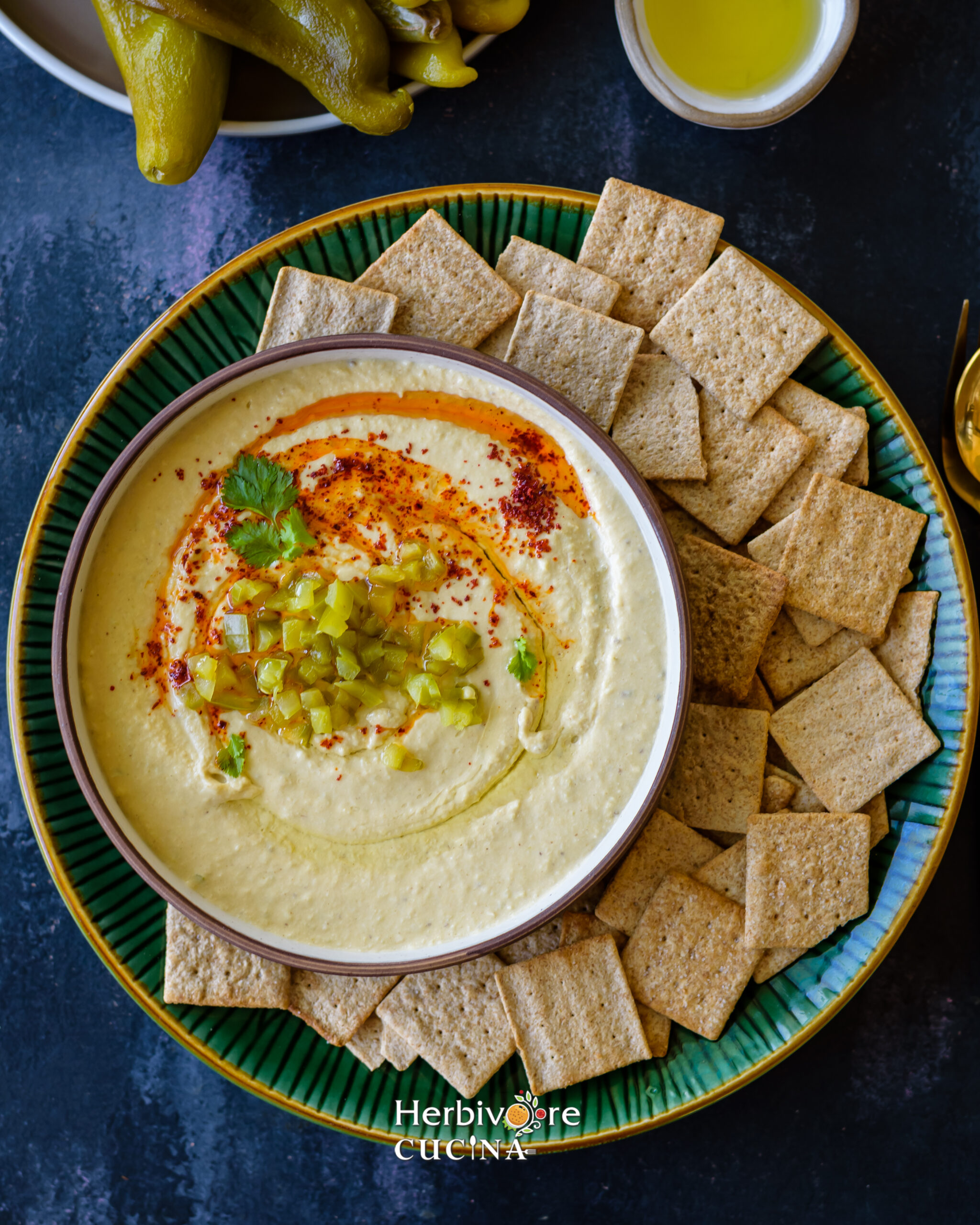 A brown bowl filled with Hatch Chile Hummus topped with oil and paprika and served with crackers in a green plate. 