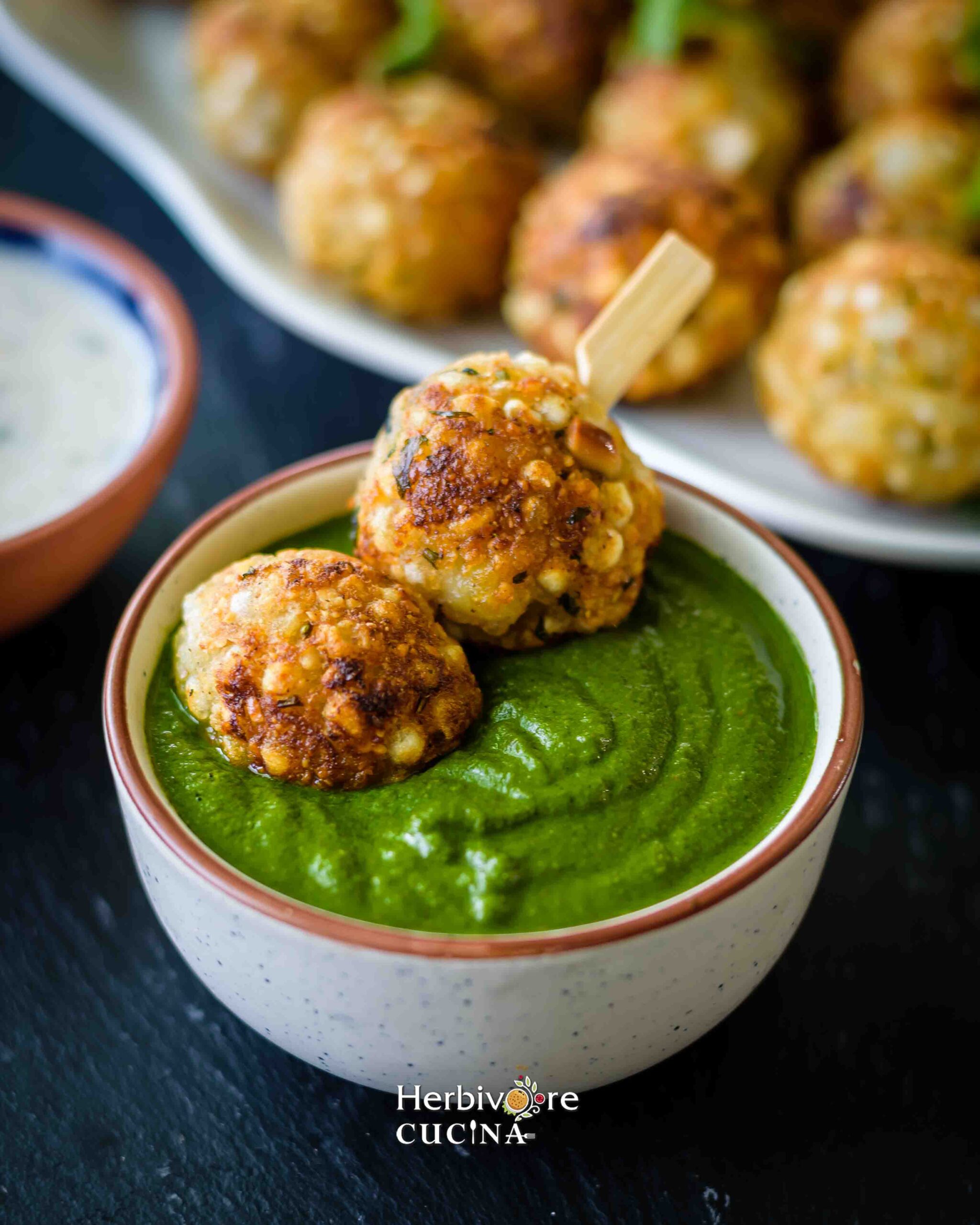 A bowl with cilantro chutney and two sabudana appe in a toothpick with more vadas in the background. 
