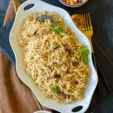 White oval plate with Parsi style brown rice topped with spices, onions and cilantro served on a black board with a brown napkin by the side.