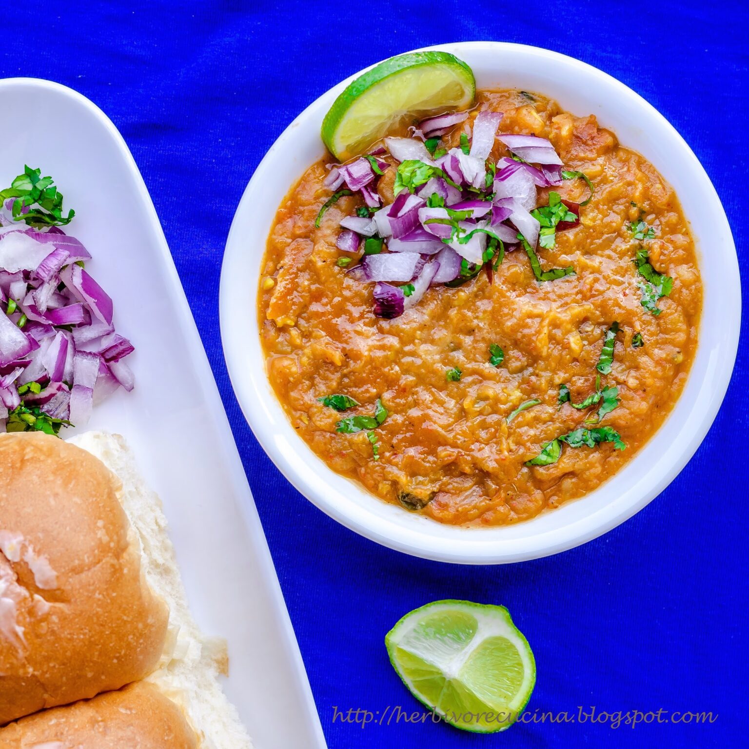 Pav Bhaji served in white bowl; topped with onions, cilantro and lemon with pav on the side. 