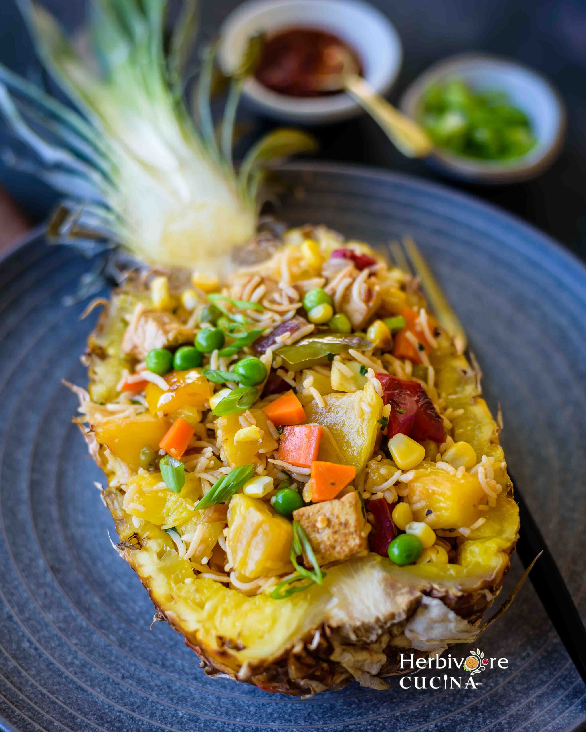Side view of half cut pineapple filled with pineapple fried rice on a gray plate with bowls of sauce and scallions on the side. 