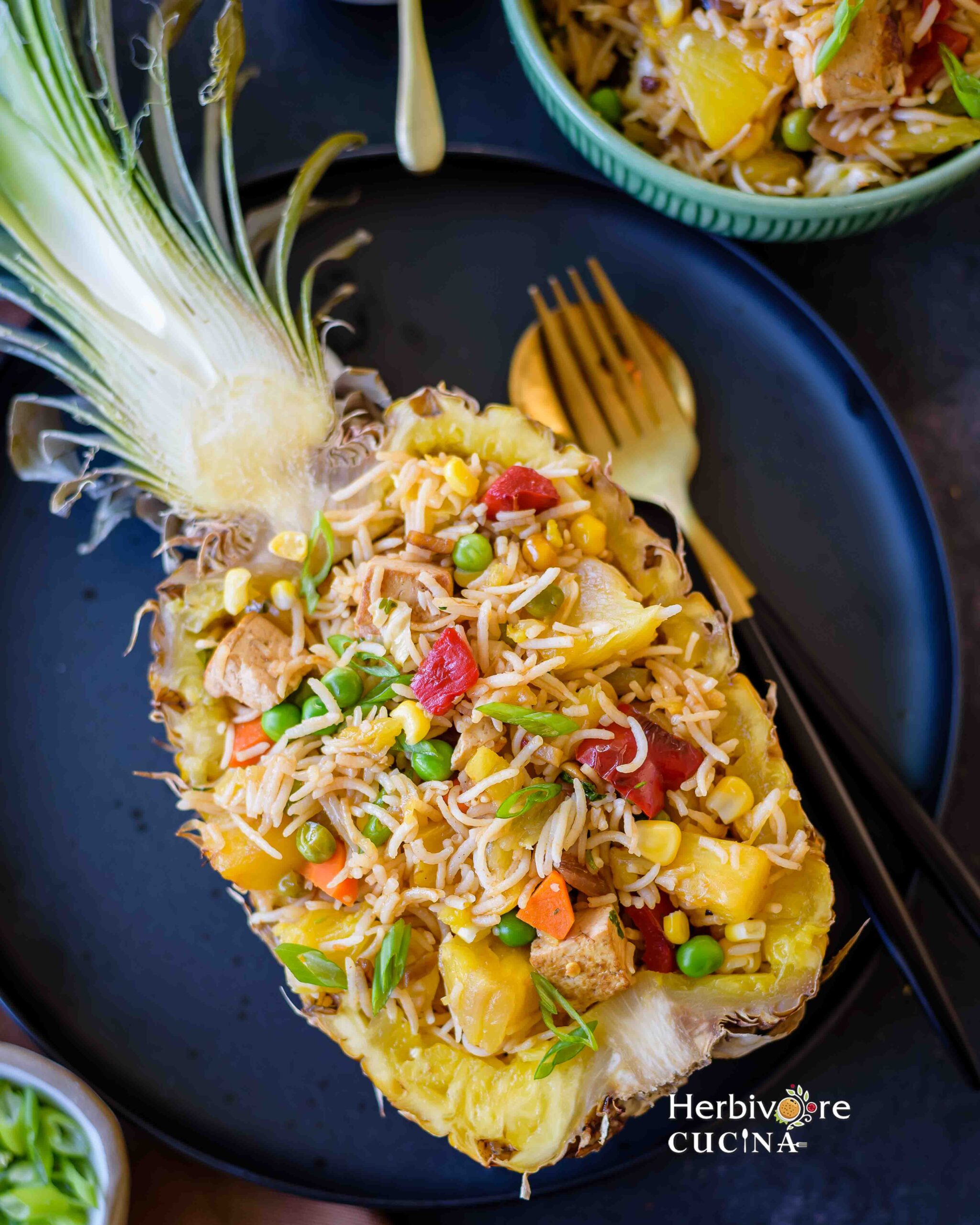 Thai Pineapple Fried Rice served in a sliced pineapple boat on a black plate and some more rice on the side with a fork. 
