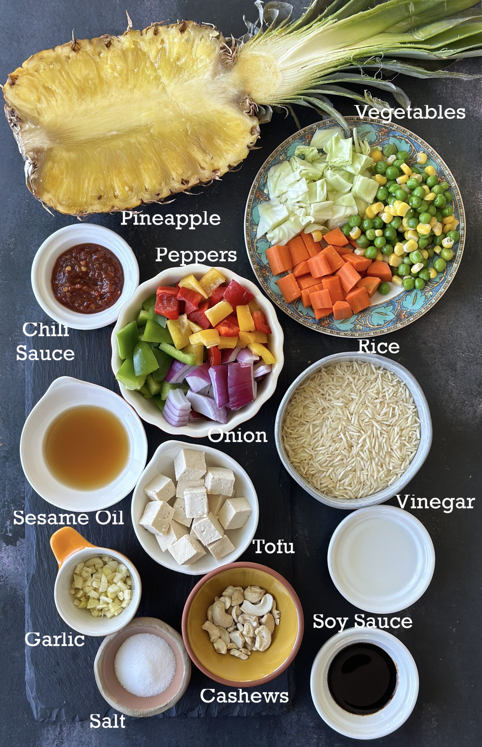 Ingredients for pineapple fried rice; pineapple, rice, vegetables and seasonings arranged on a black surface. 