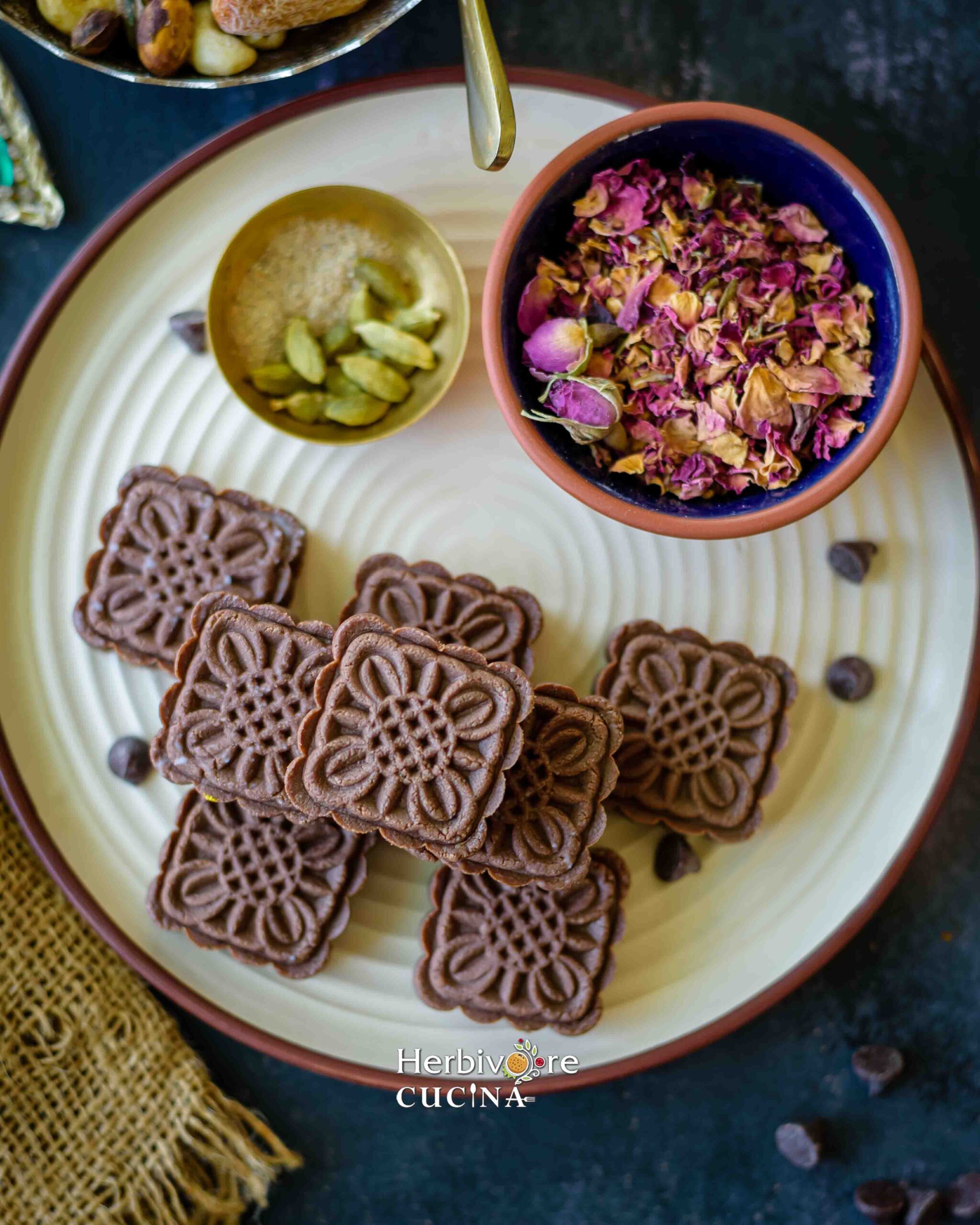 Chocolate peda on a white plate with rose petals and elaichi on the side. 