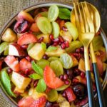 Fruit Chaat in a black and white bowl with a golden fork and spoon by the side and mint leaves around it.