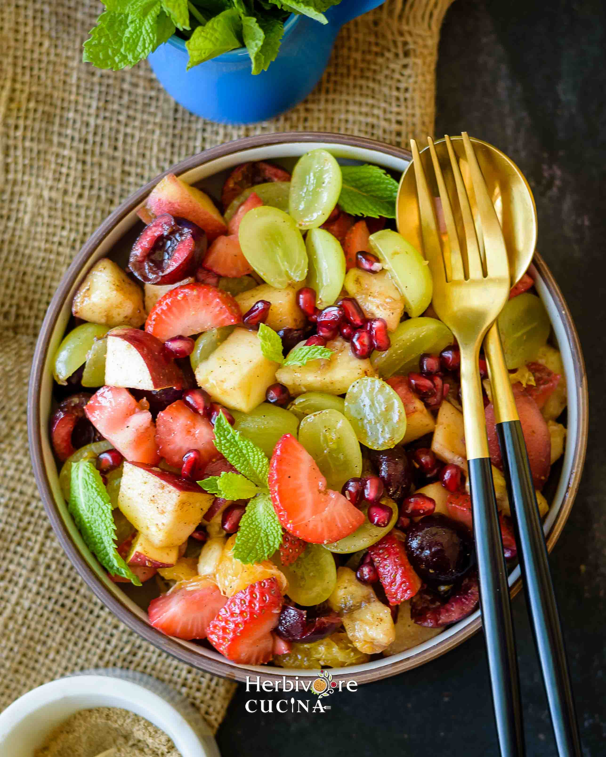 Fruit Chaat in a black and white bowl with a golden fork and spoon by the side and mint leaves around it. 
