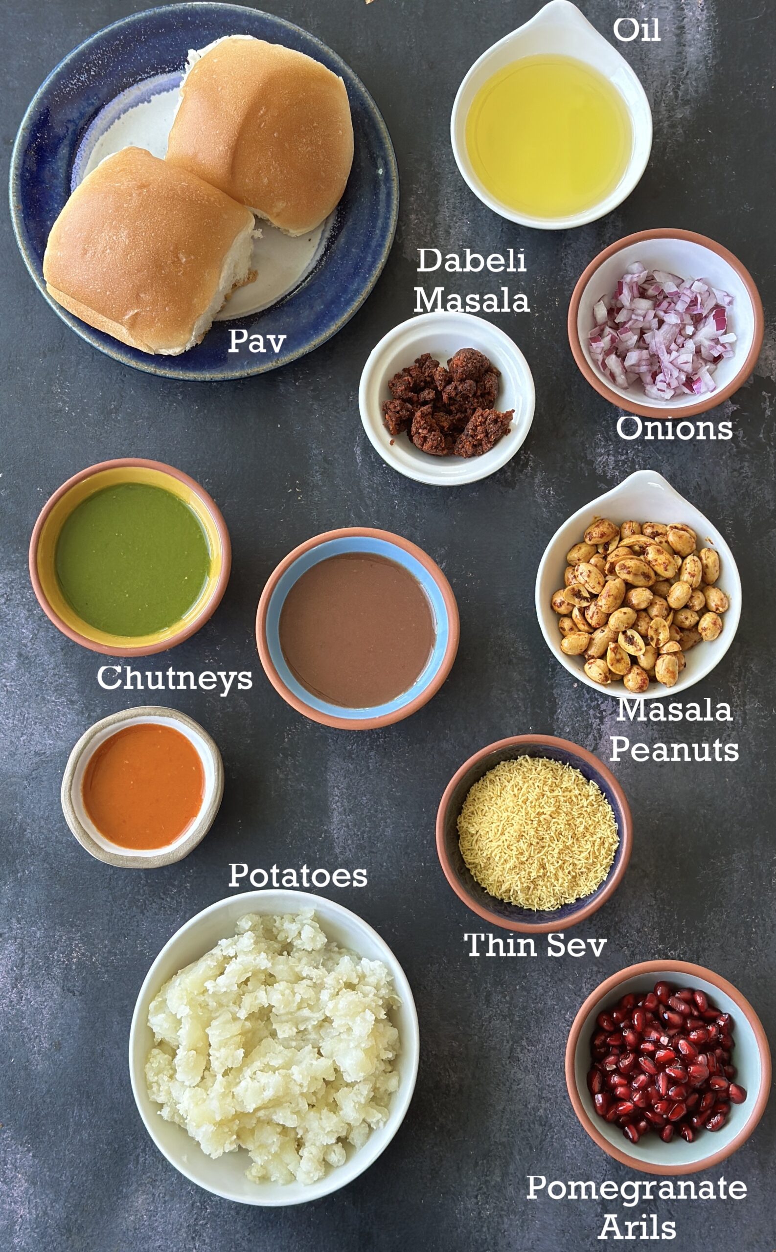 Ingredients for Kutchi Dabeli; bread, potatoes, seasonings and toppings on a dark background. 