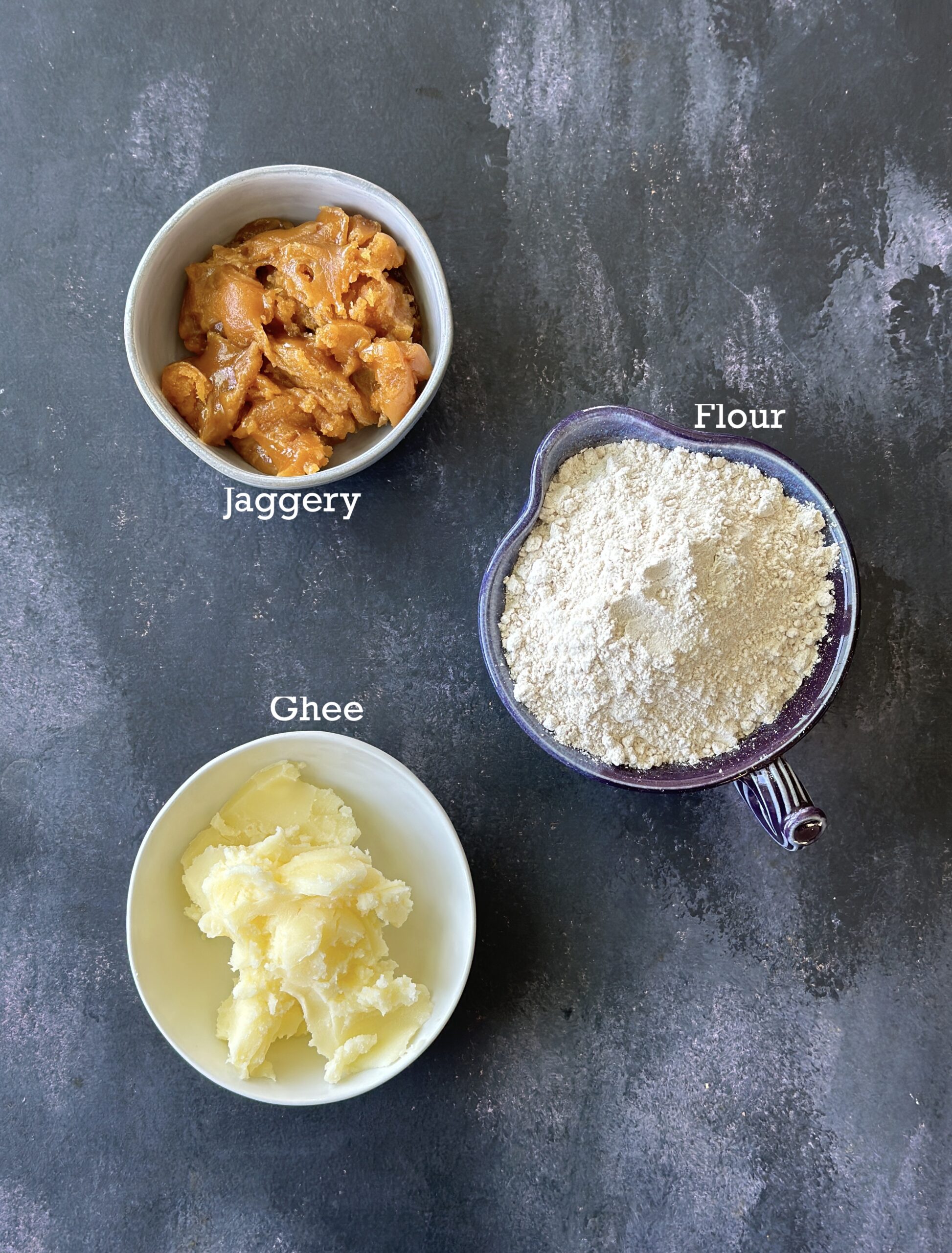 Ingredients for sukhdi; jaggery, flour and ghee in bowls on a black board. 