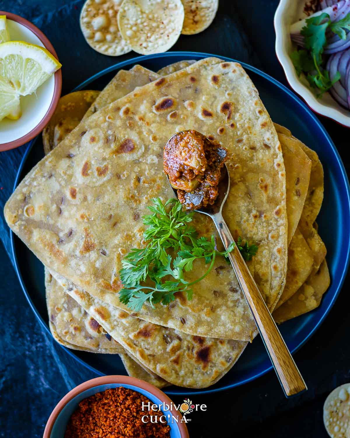 Triangle parathas stacked in a blue plate with some Indian pickle and cilantro placed on top.