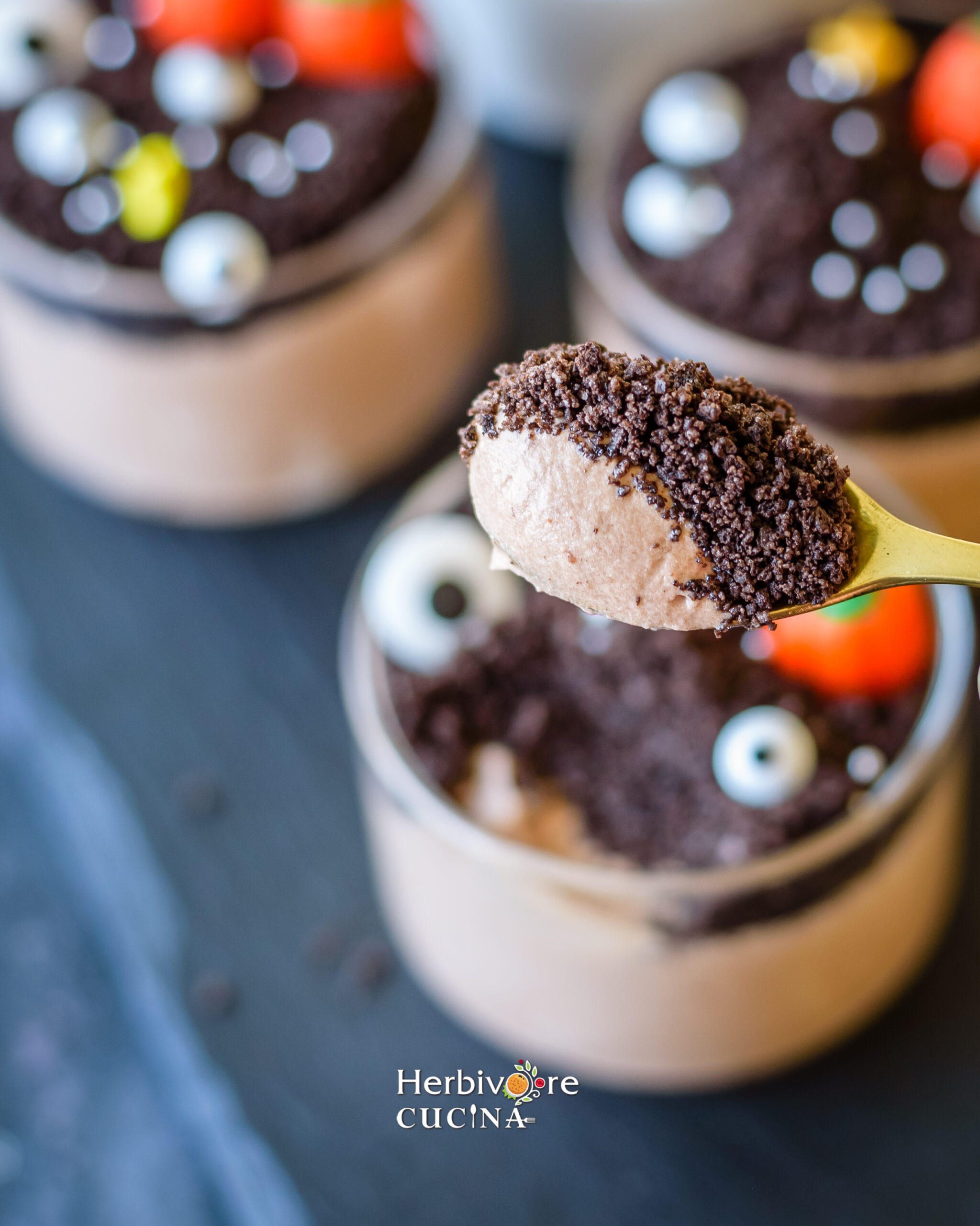 A spoon with chocolate mousse and crushed oreo with Halloween themed mousse in the background.