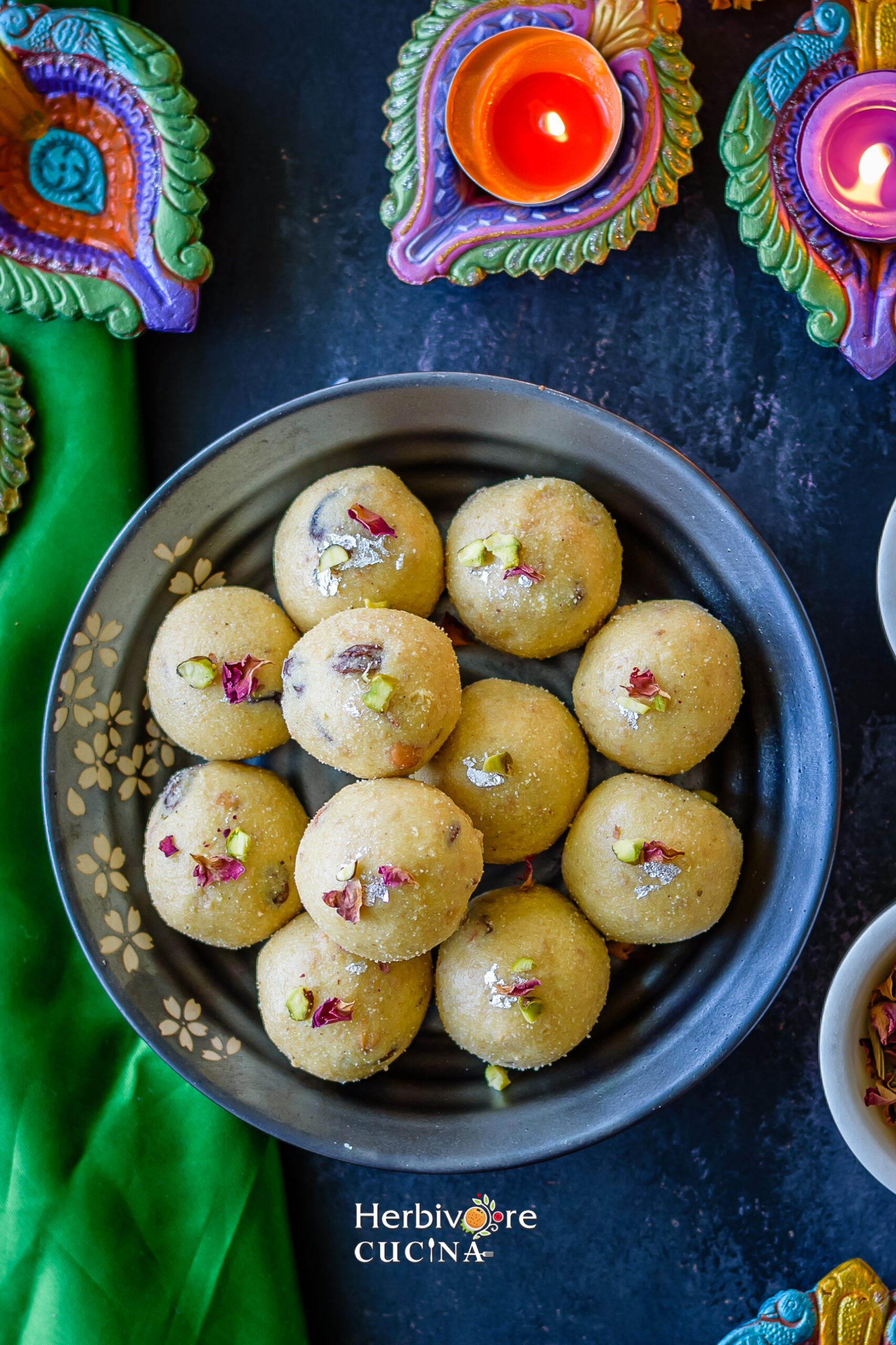 A dozen rava ladoo placed on a plate and topped with rose petals and nuts with lit diyas on the side. 