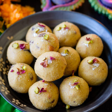 Rava Ladoo in a plate topped with dried rose petals and silver foil with diyas in the background.