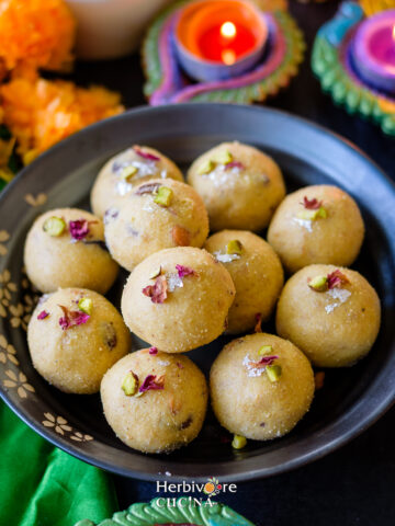 Rava Ladoo in a plate topped with dried rose petals and silver foil with diyas in the background.