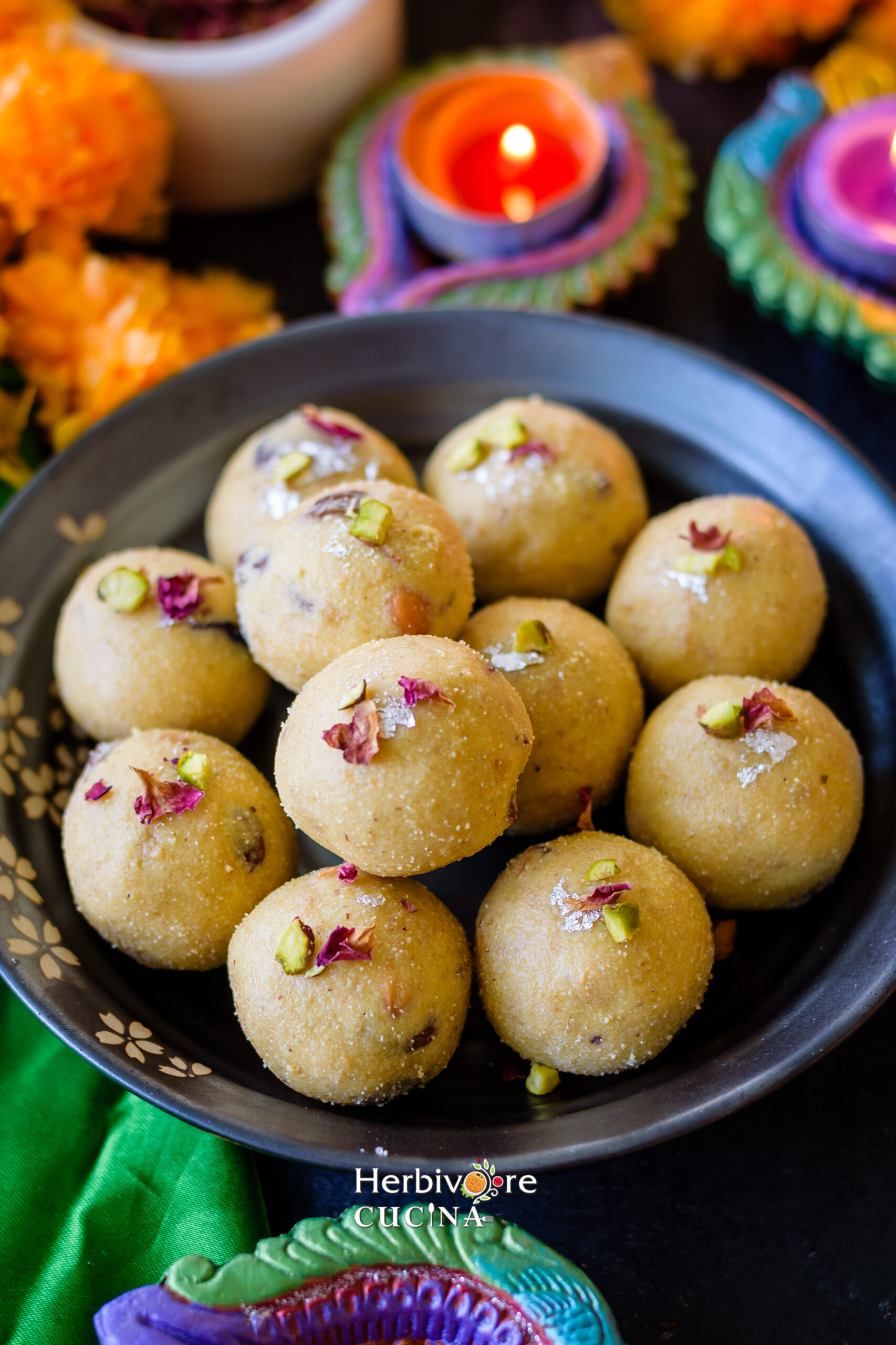 Rava Ladoo in a plate topped with dried rose petals and silver foil with diyas in the background. 