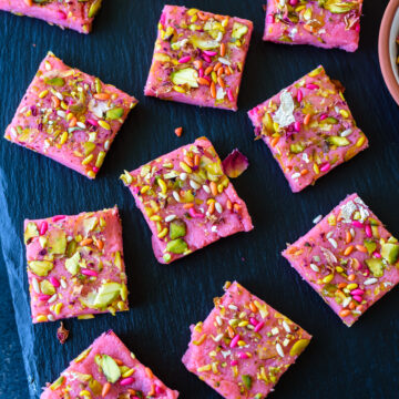 A slate board with a dozen pieces of rose kalakand topped with pistachios and fennel seeds.