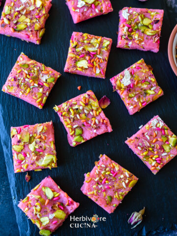 A slate board with a dozen pieces of rose kalakand topped with pistachios and fennel seeds.