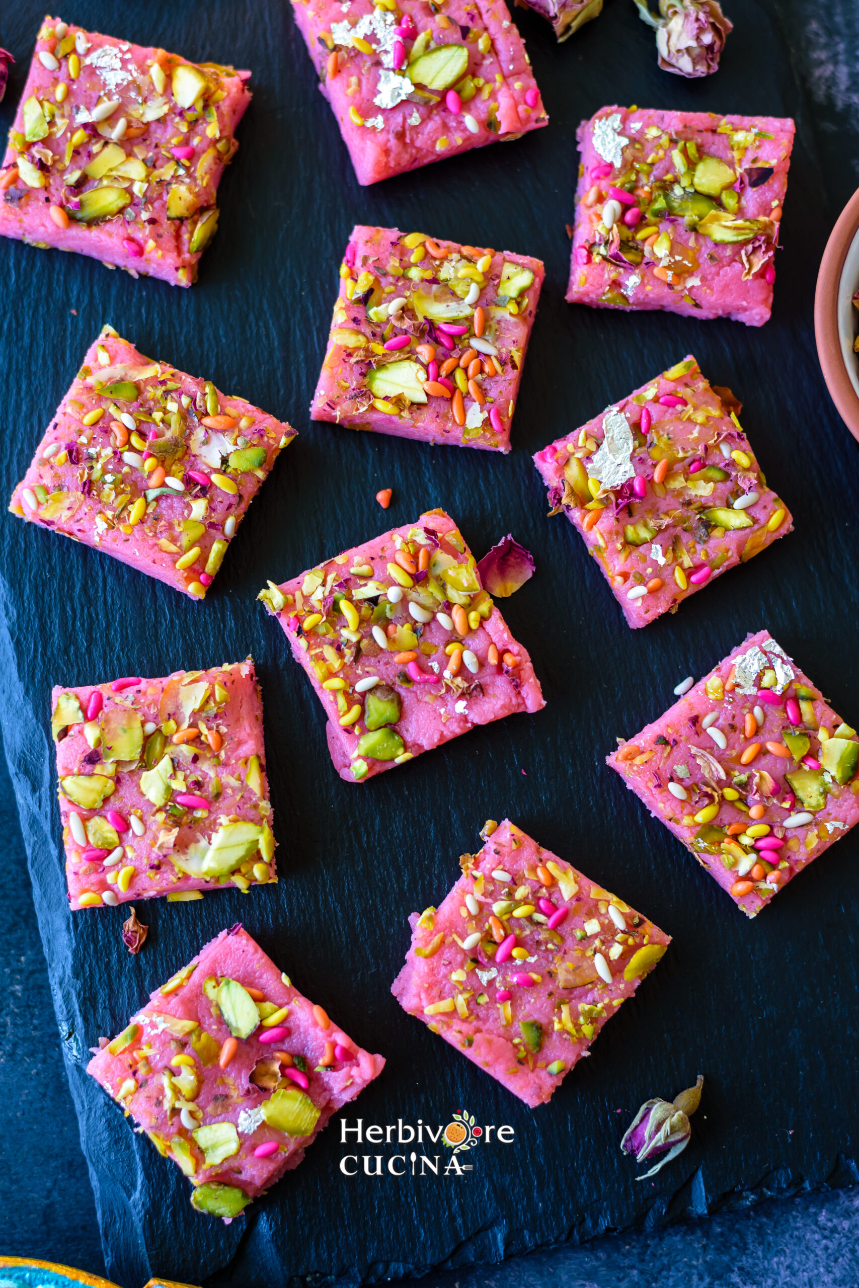 A slate board with a dozen pieces of rose kalakand topped with pistachios and fennel seeds. 