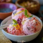 A bowl of rose falooda ice cream topped with roe syrup, sev and takmaria served with a spoon.