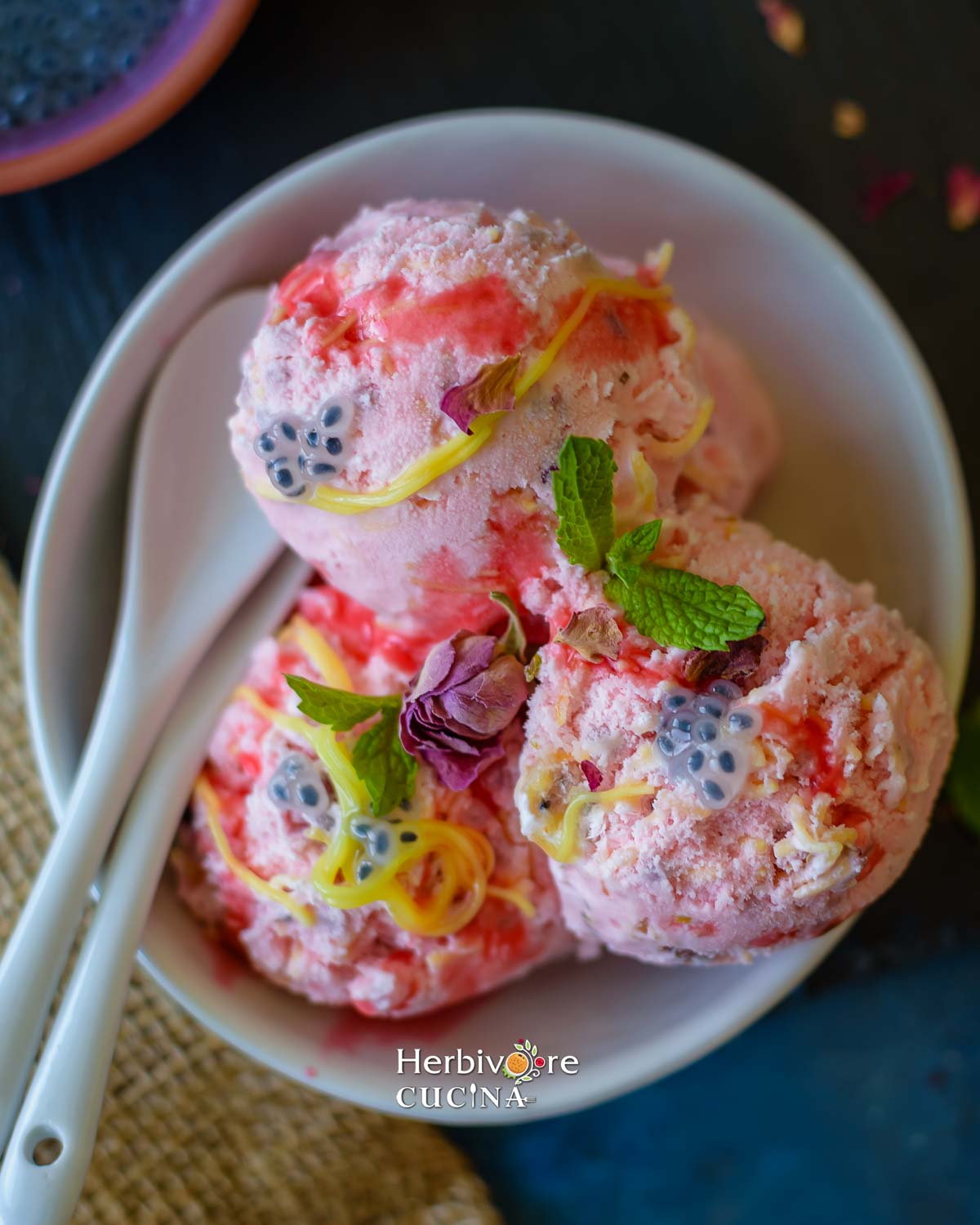 A bowl with rose falooda ice cream; topped with basil seeds, rose syrup and sev; with a spoon on the side. 