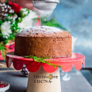 A Christmas fruit cake on a red cake pedestal with a sprinkle of sugar above.