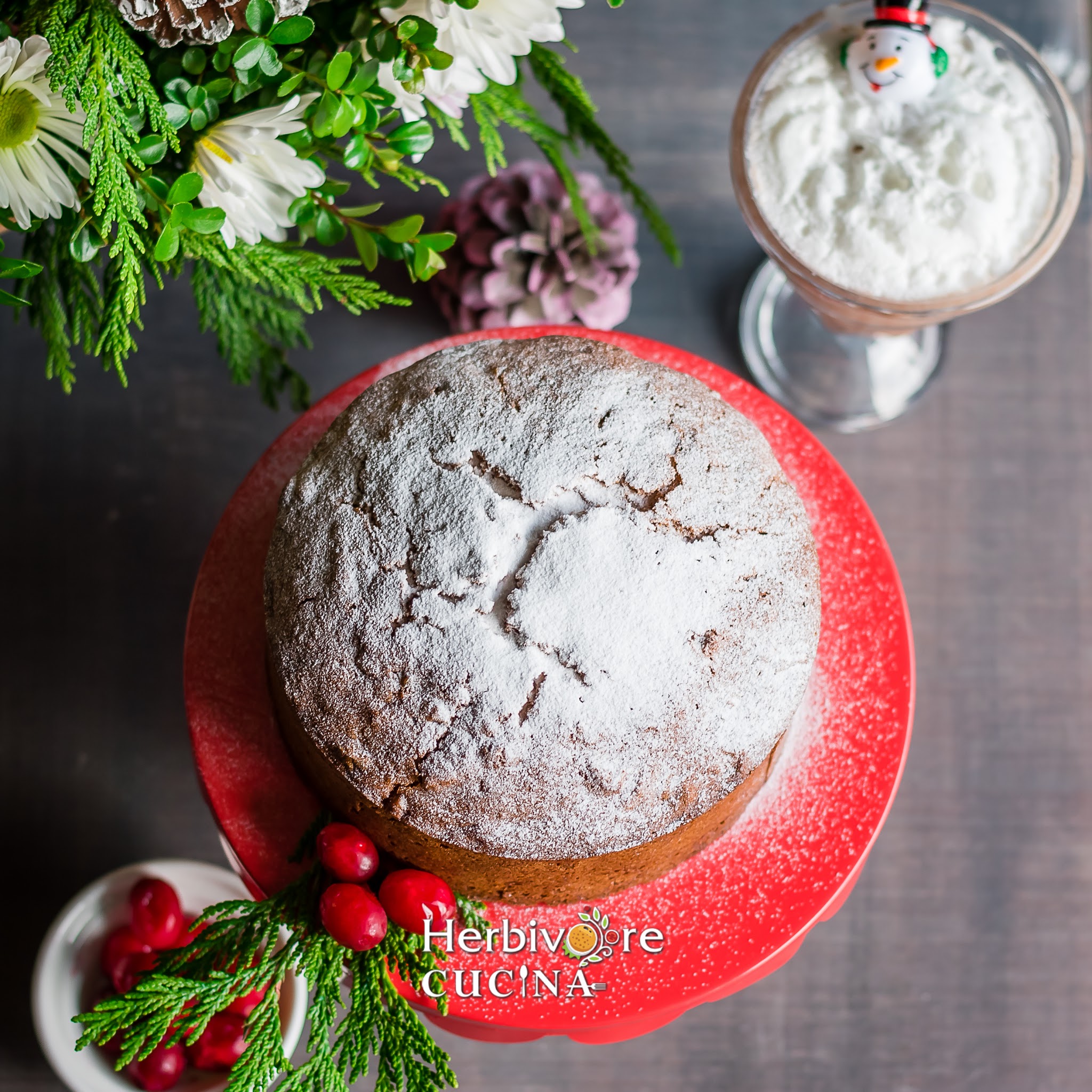 An eggless Christmas cake topped with powdered sugar and some flowers and hot chocolate on the side. 