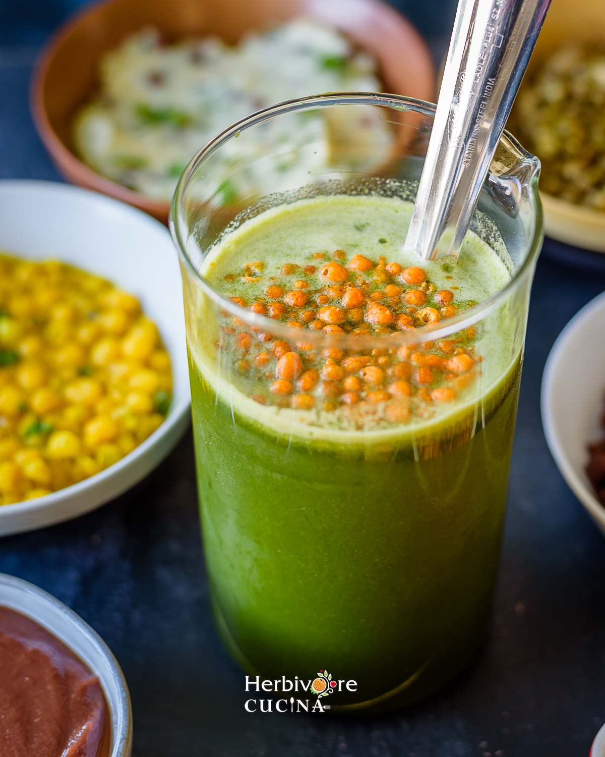A glass container filled with spicy pani and topped with boondi with other pani puri ingredients around it. 