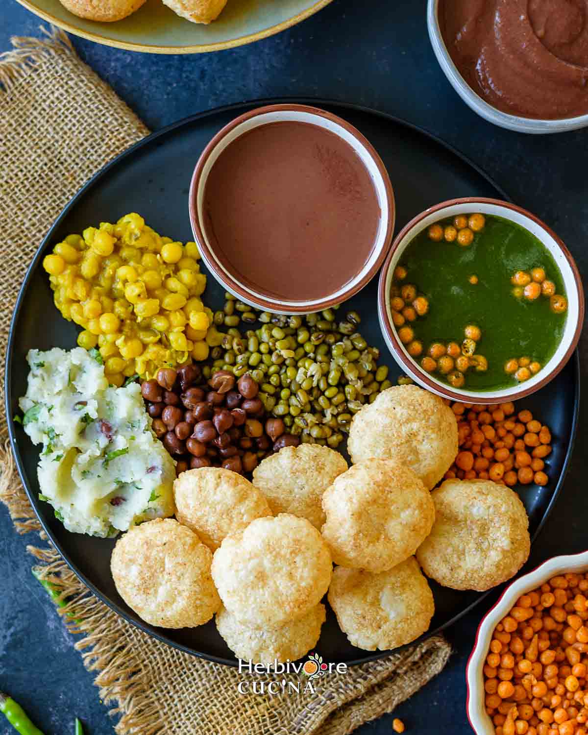 A black plate filled with puri, stuffings, spicy pani and sweet chutney in small bowls. 