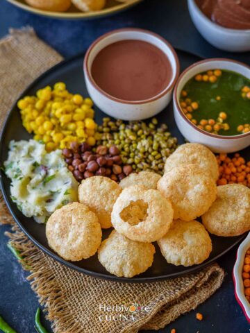 A plate of pani puri ingredients in a black plate with puri, chutney, spicy pani and add-ins.