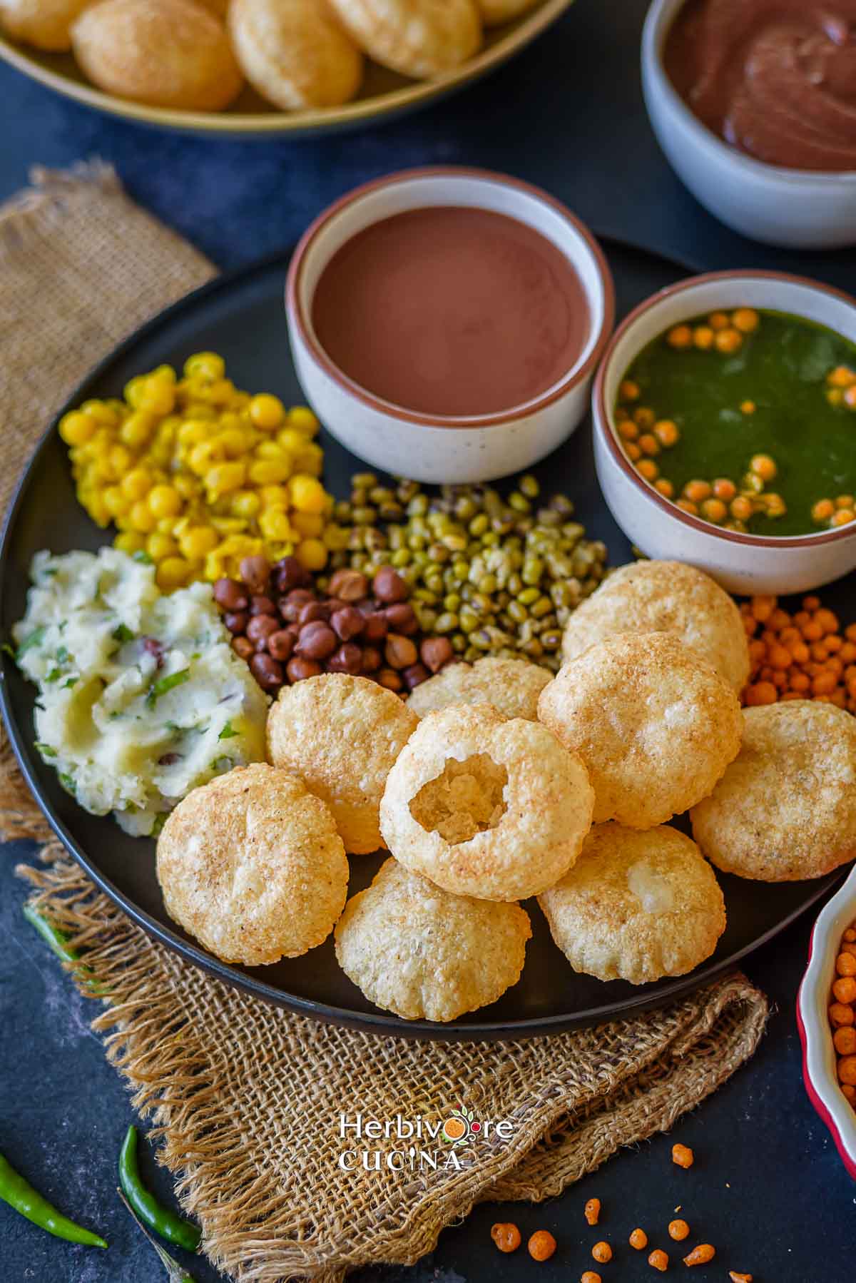 A plate of pani puri ingredients in a black plate with puri, chutney, spicy pani and add-ins. 