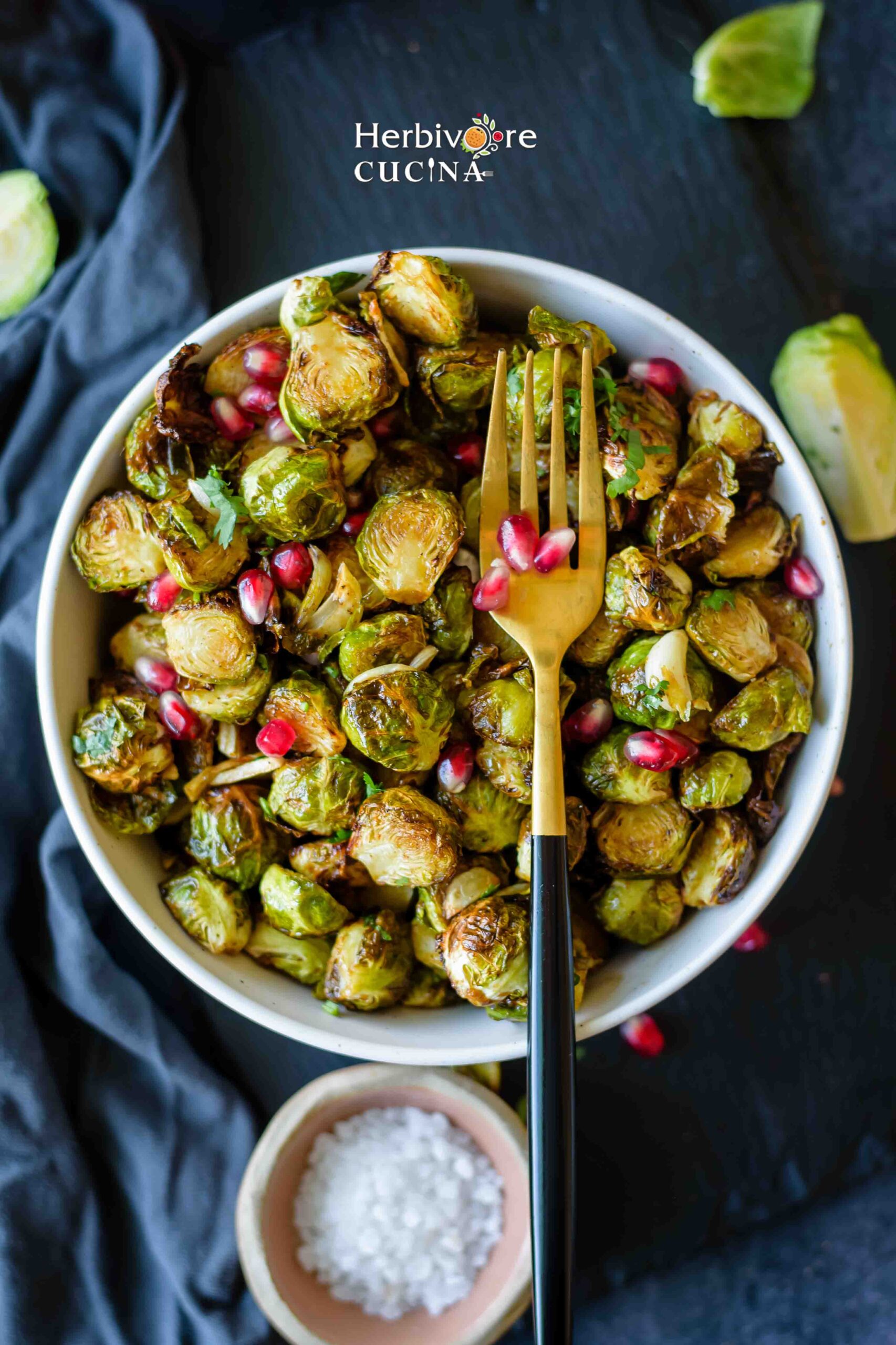 Air Fryer Brussels Sprouts in a bowl topped with pomegranate arils and a small bowl of salt beside it. 