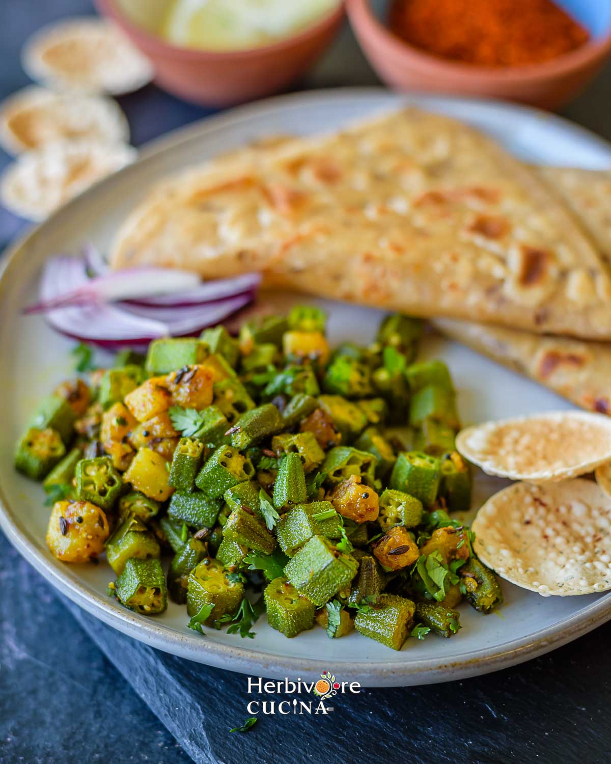 Okra potato sabzi with parathas in a plate and some papad and onions on the side. 
