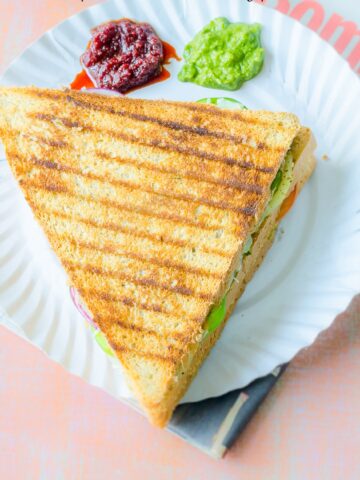 Triangular grilled sandwich on a paper plate with cilantro chutney and hot sauce on the side.