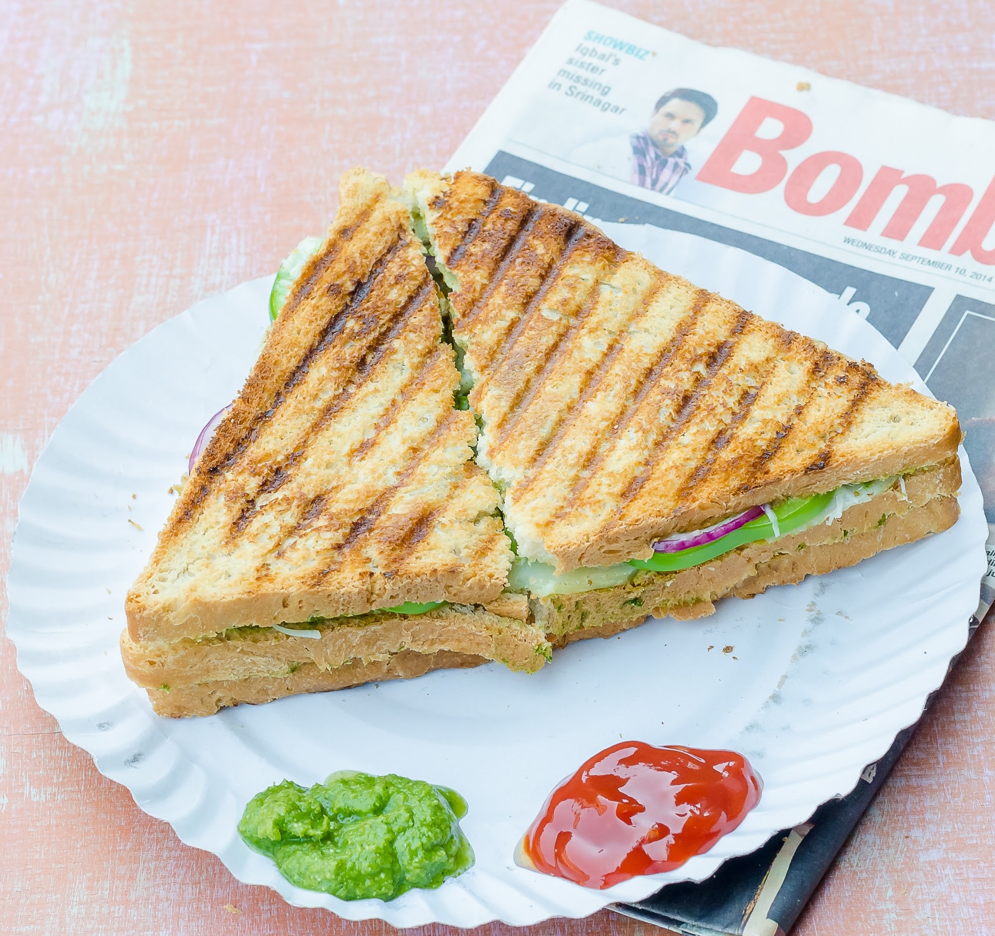 Sliced Bombay grilled sandwich on a plate with cilantro chutney and sauce on the side on a copy of Bombay Times. 