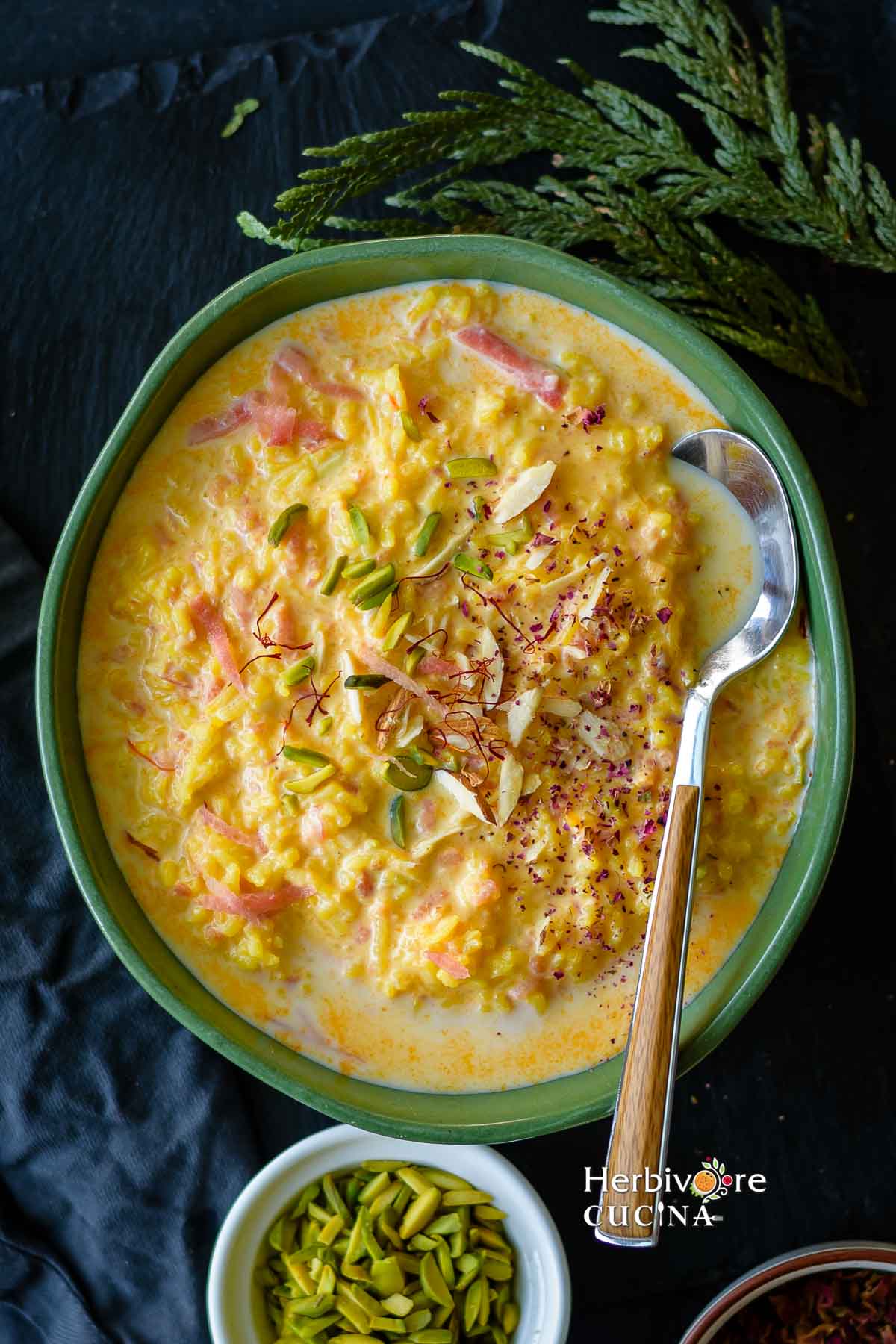A green bowl filled with carrot kheer topped with nuts, rose petals and saffron with a spoon on the side. 