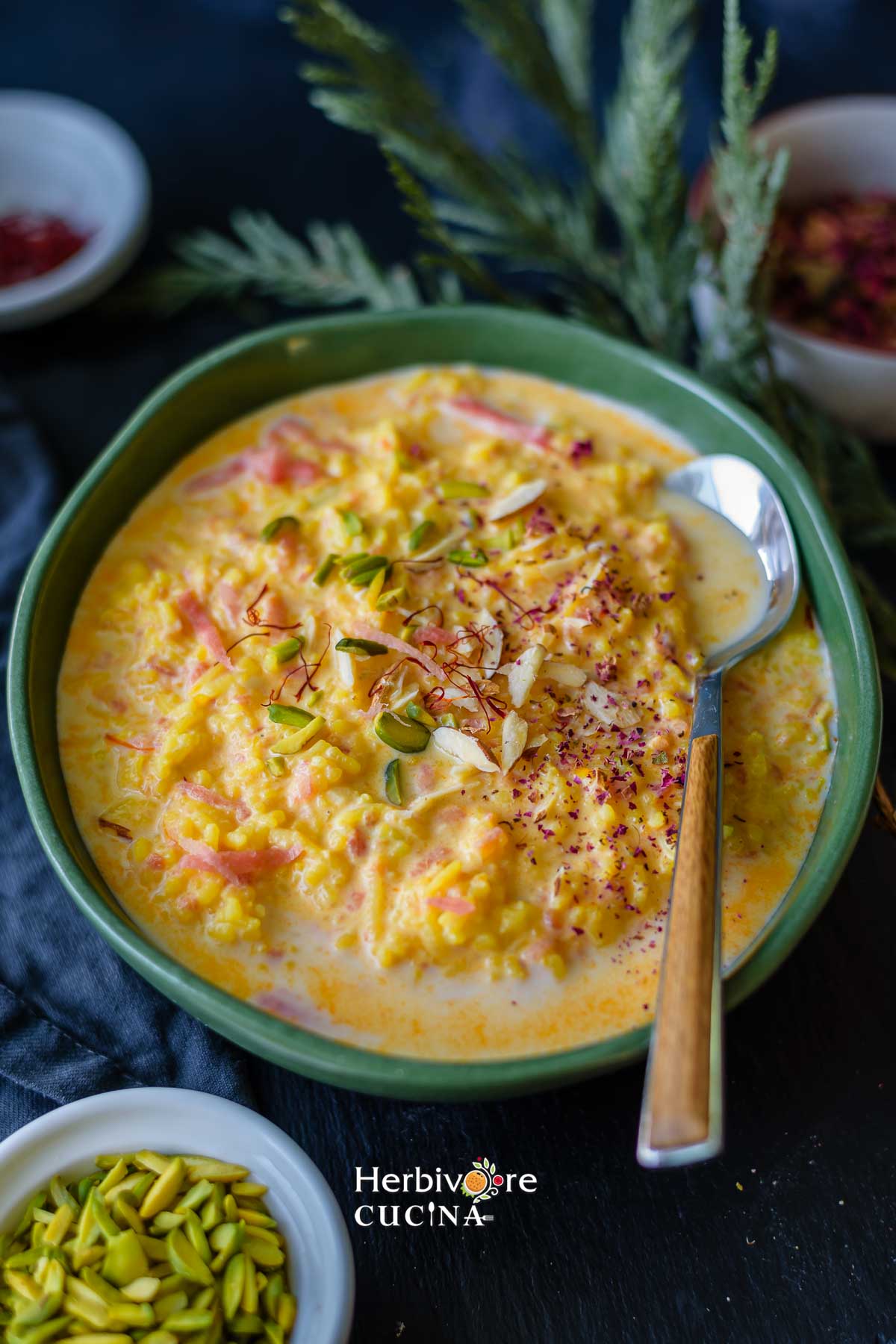 An oval bowl with carrot kheer; topped with nuts, rose petals and saffron with a spoon on the side and nuts in small bowls. 