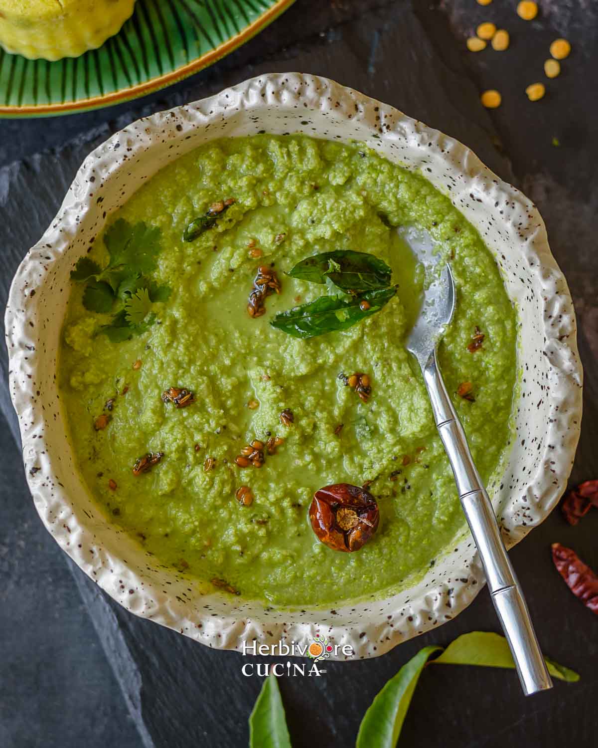 A white bowl with coconut cilantro chutney with tempering and a spoon on the side. 