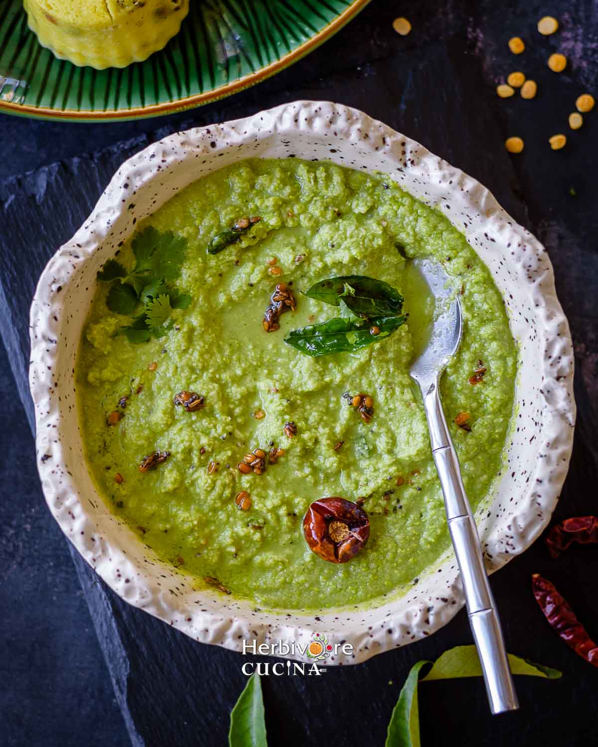 Coconut cilantro chutney in a white bowl with tempering on top and a spoon on the side. 