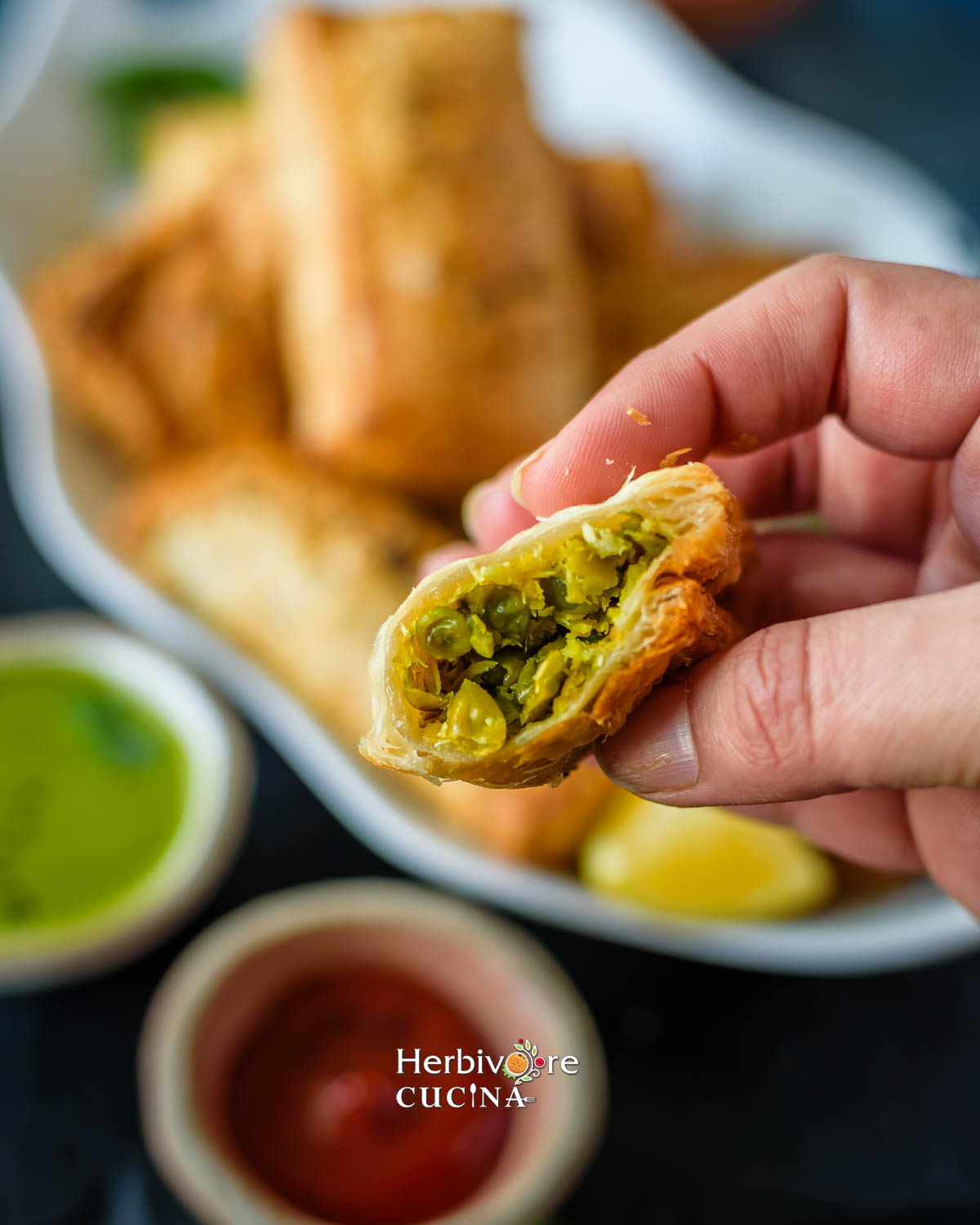 Holding a cut green peas kachori puff showing the filling and puff layers with some ketchup and chutney on the side. 