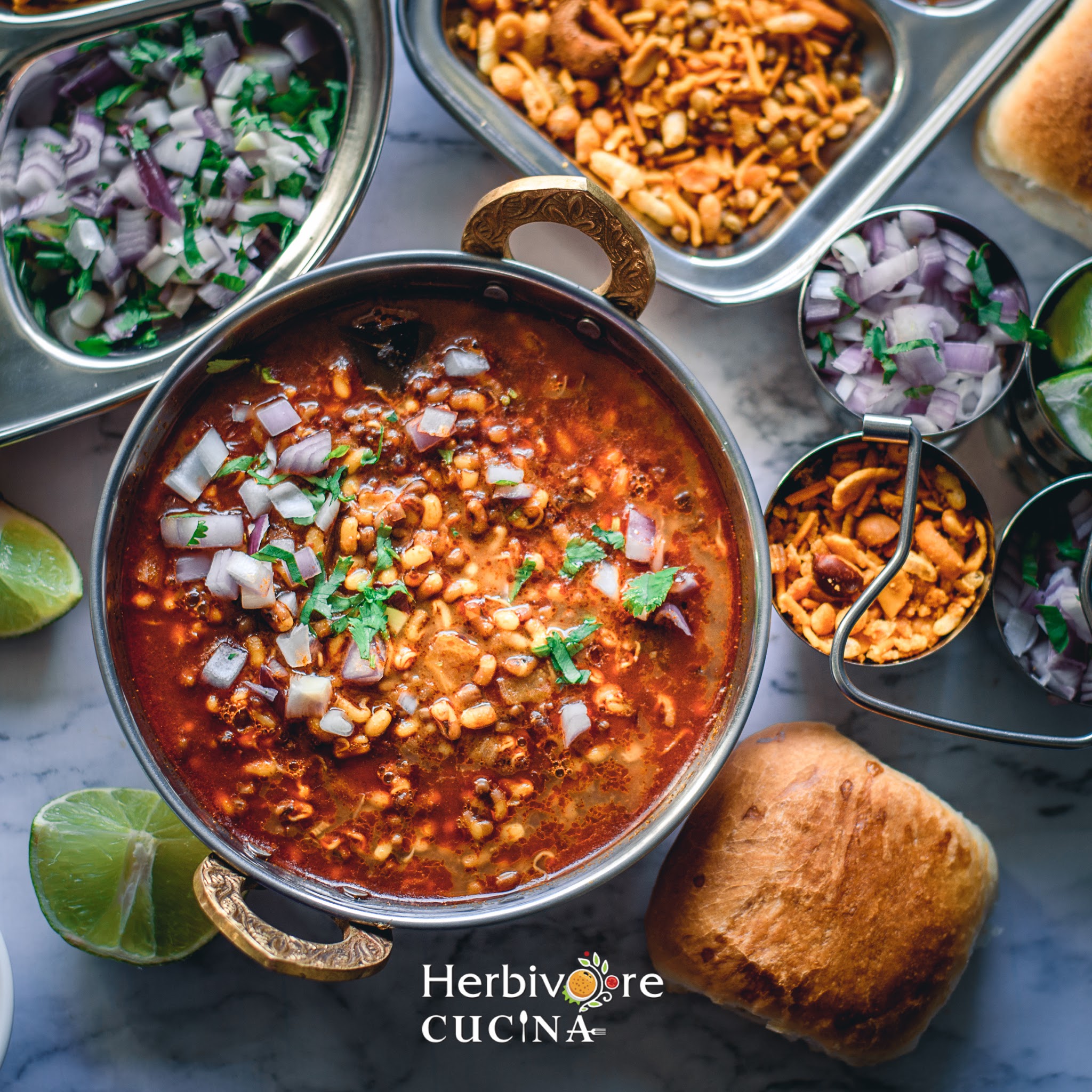 A metal bowl filled with matki misal, topped with onions and served with different add-ins and pav bread. 