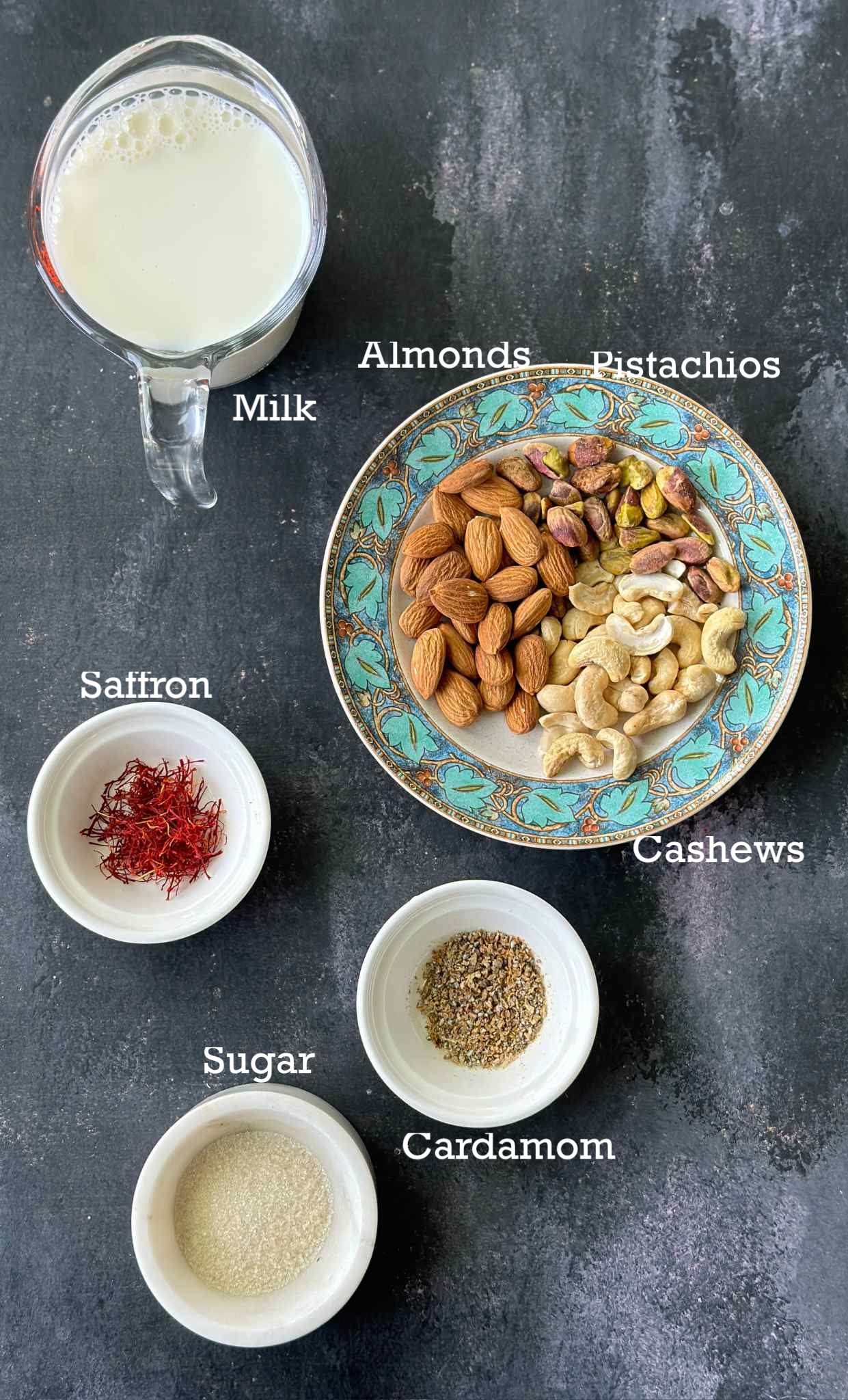 Labelled ingredients to make masala milk in small bowls on a dark surface. 