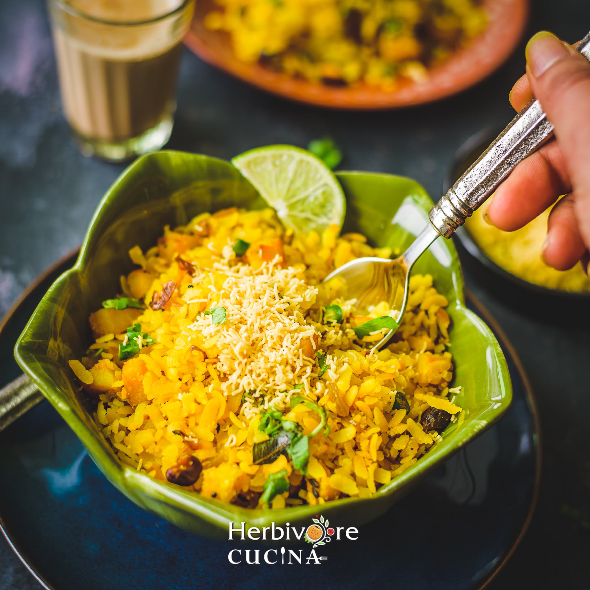 A spoon held over a bowl filled with batata poha with a lemon slice and sev on it. 