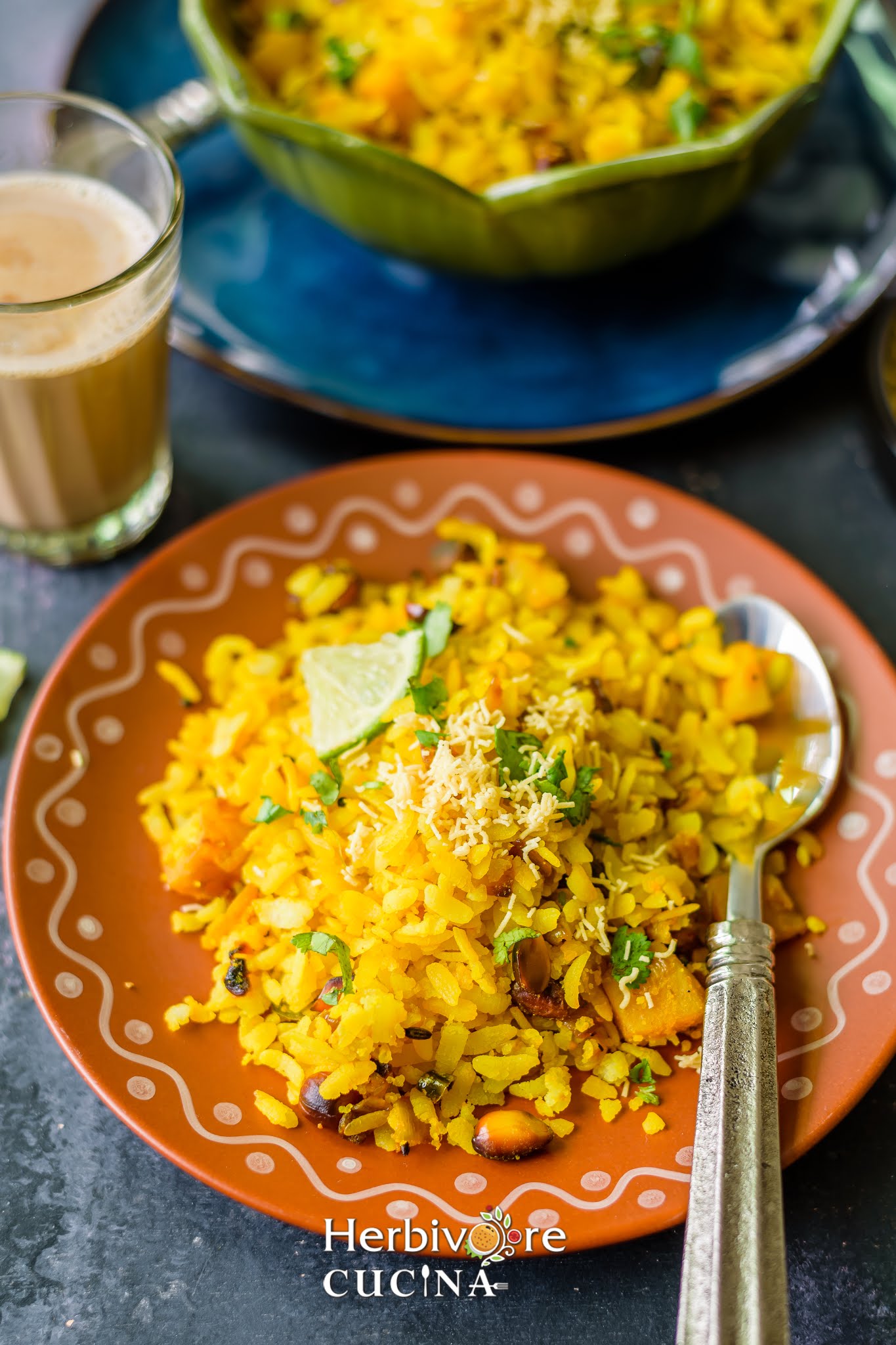 A brown plate with batata poha and. spoon beside it; topped with sev and a lemon wedge. 