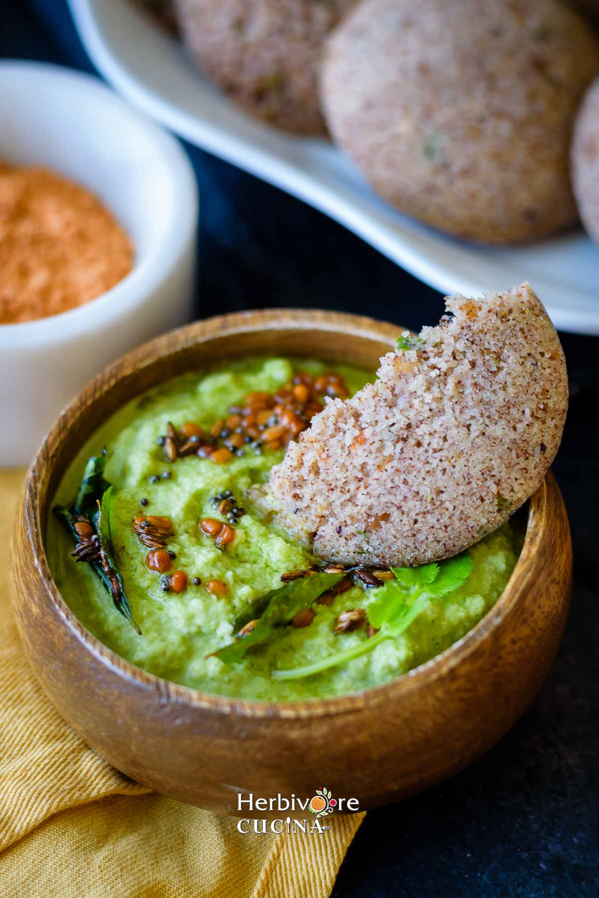 A bowl of cilantro coconut chutney with a half cut ragi rava idli dipped in it. 