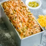 A tin bread pan with a loaf of baked jalapeño and cheddar bread with more toppings on the side.