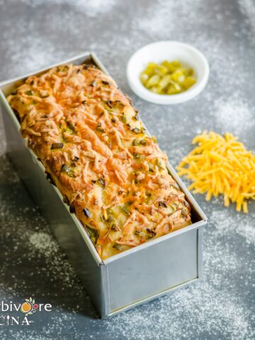 A tin bread pan with a loaf of baked jalapeño and cheddar bread with more toppings on the side.
