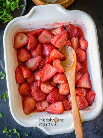 Roasted Strawberries in a white platter with a spoon on the side and leaves on the side.