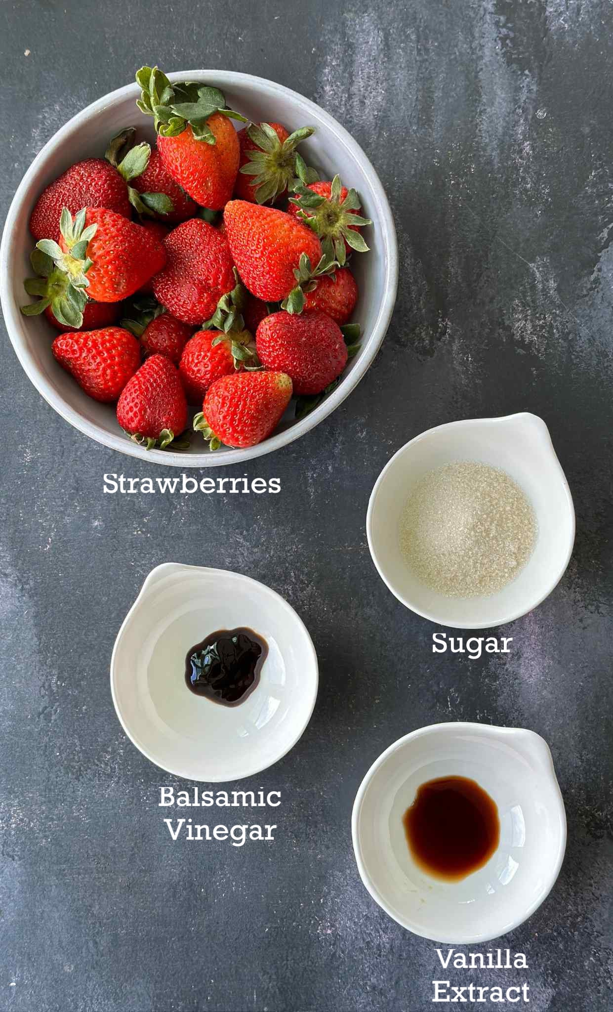Ingredients for roasted strawberries in small bowls on a dark surface. 