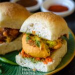 Vada pav placed in a green plate with bowls of chutney placed behind it.