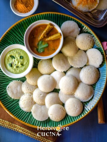 Instant Pot idli in a green plate with chutney and sambar around it in small bowls.