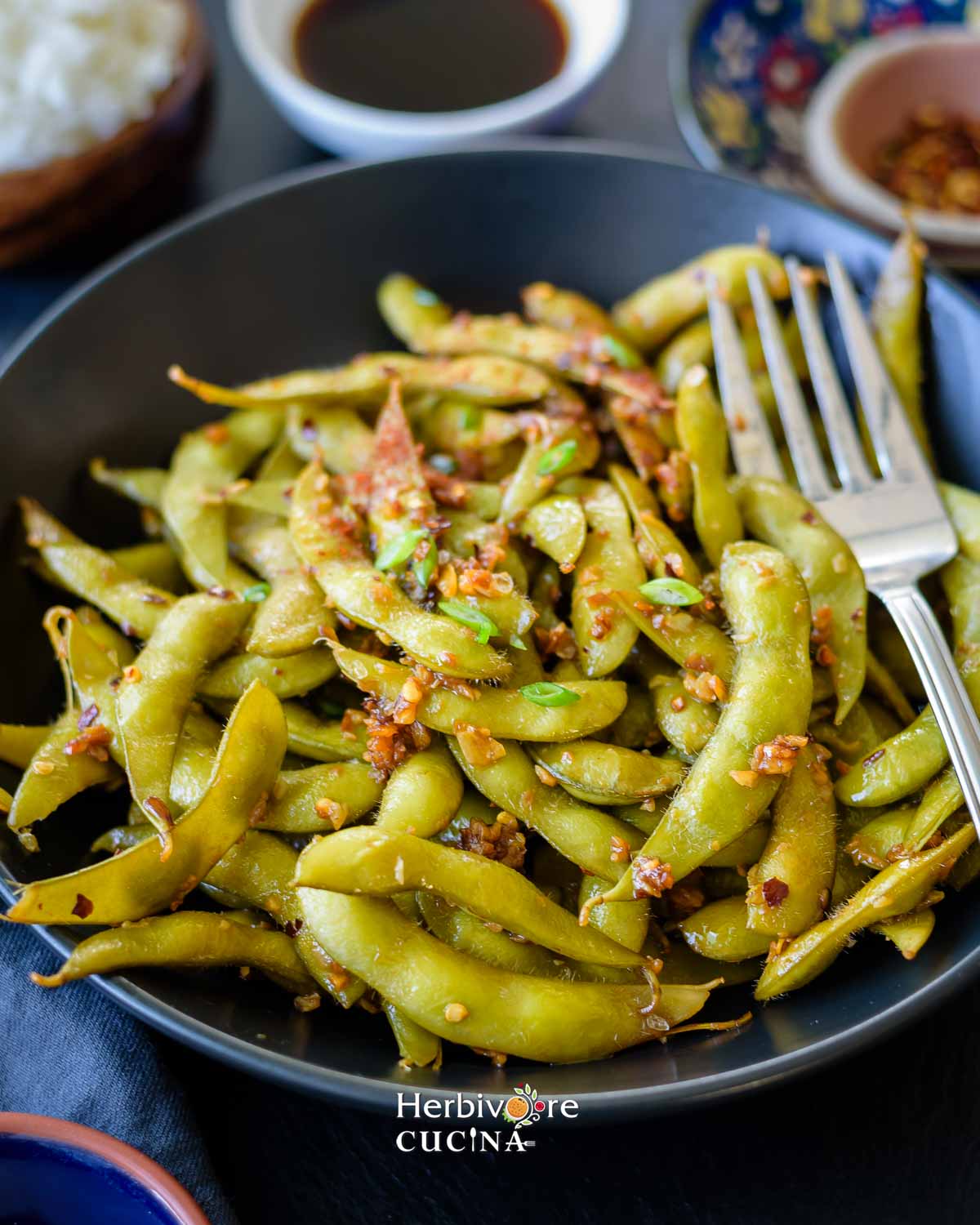 A bowl of garlic edamame with a fork on the side of the bowl with more seasonings on the side. 